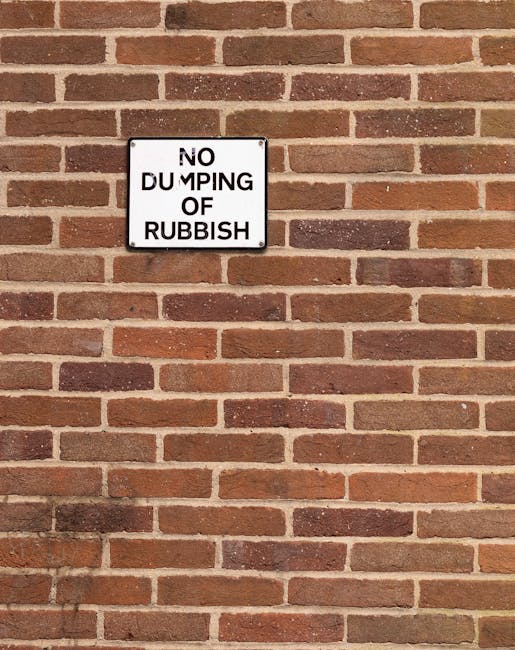 A rectangular metal sign with a white background and black border mounted on a red brick wall. The sign displays the message 'NO DUMPING OF RUBBISH' in bold black capital letters, with the words arranged in four lines. The brick wall has a uniform pattern with reddish-brown and dark brown bricks, some with slight variations in texture and shade. The mortar between the bricks is light grey, creating a clear contrast with the bricks. The overall scene depicts an outdoor wall, possibly at the side or back of a building, with natural lighting casting minimal shadows and highlighting the texture of the brickwork and the sign, reflecting a setting related to waste management or property boundary markings.