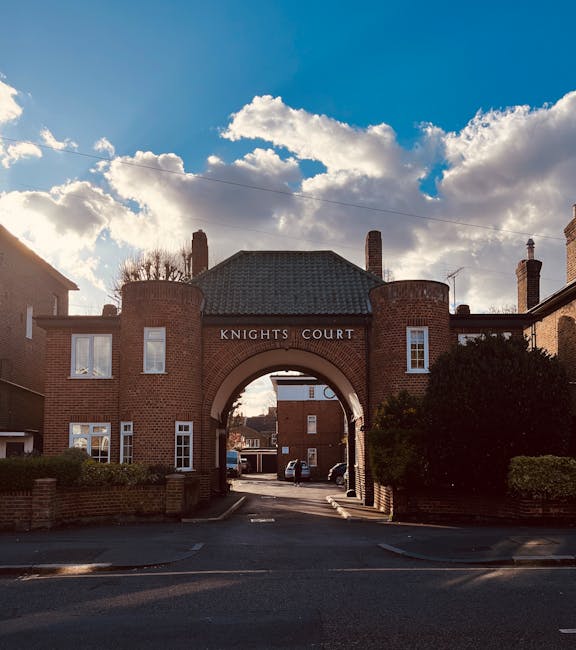 A brick archway with the inscription 'KNIGHTS COURT' spans a narrow driveway, forming the entrance to a residential complex, with two rounded brick towers on either side and small, white-framed windows. The top of the arch is topped with a pitched, dark tiled roof. Behind the arch, parked cars are visible along a quiet street lined with brick buildings. The sky above is partly cloudy with large, white clouds and patches of blue, casting soft light over the scene. The surrounding area includes trimmed hedges and brick walls, and the environment appears clean and well-maintained. This setting exemplifies a typical private residential entrance, which may be relevant when considering alternative waste collection or on-site clearance options, as promoted by independent rubbish removal services in Kingston upon Thames.