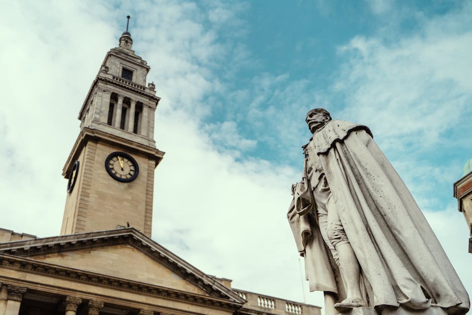 A historic clock tower with a beige stone façade and a black clock face displaying the time, set against a partly cloudy sky with blue and white hues. In the foreground on the right side, a detailed stone statue of a man dressed in period clothing, including a long coat and a hat, stands on a pedestal. The statue's surface appears weathered with visible texture and slight discoloration. The clock tower's upper section features a spire topped with a small decorative element. The building beneath the clock tower has classical architectural elements such as a triangular pediment supported by columns. The scene captures a blend of architectural and sculptural features, suggesting a historic public square or town centre setting, which aligns with the context of public or private waste management services in an urban environment, although no rubbish or waste items are visible in the image.