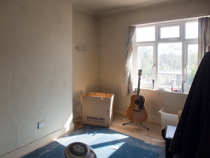 Inside a room with beige painted walls and a large double-glazed window allowing natural light, there is a cardboard box labeled 'Shurgard Self-Storage' placed on the wooden floor near the corner, with its flaps partially open. Next to the box, a wooden acoustic guitar with a light natural finish leans against the window, its dark fretboard and tuning pegs visible. The window frames a view of a suburban street with trees and a house in the background, partially obscured by light-colored curtains hanging on either side. On the floor, a small round robot vacuum cleaner is situated in the foreground, casting a shadow on a blue rug. Nearby, a plastic storage bin is positioned against the wall, with a black coat or fabric draped over an object on the right side of the image. The overall scene suggests a modest, inhabited room in the process of tidying or clearing, representative of an estate or home clearance service environment, as managed by rubbishremovalkingstonuponthames.co.uk.