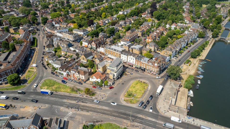 An aerial view of a small town showing a dense arrangement of medium-height residential and commercial buildings with a variety of roof styles, including flat and pitched tiles in shades of grey and brown. The streets are busy with cars, trucks, and buses, some parked along the curbs and others in motion. In the lower part of the image, a main road runs horizontally, with a smaller roundabout and a few patches of green space adjacent to it. To the right, a waterway with a concrete embankment features several docked boats, with a paved walkway along the waterfront. The background reveals a lush, leafy suburban area with many trees and houses, extending into a densely treed landscape. The scene is well-lit with natural daylight, casting soft shadows, and conveys an urban environment that may include services like rubbish collection and waste management, as visible through the organized traffic and infrastructure supporting local waste disposal activities, consistent with a typical town setting observed from an aerial perspective.