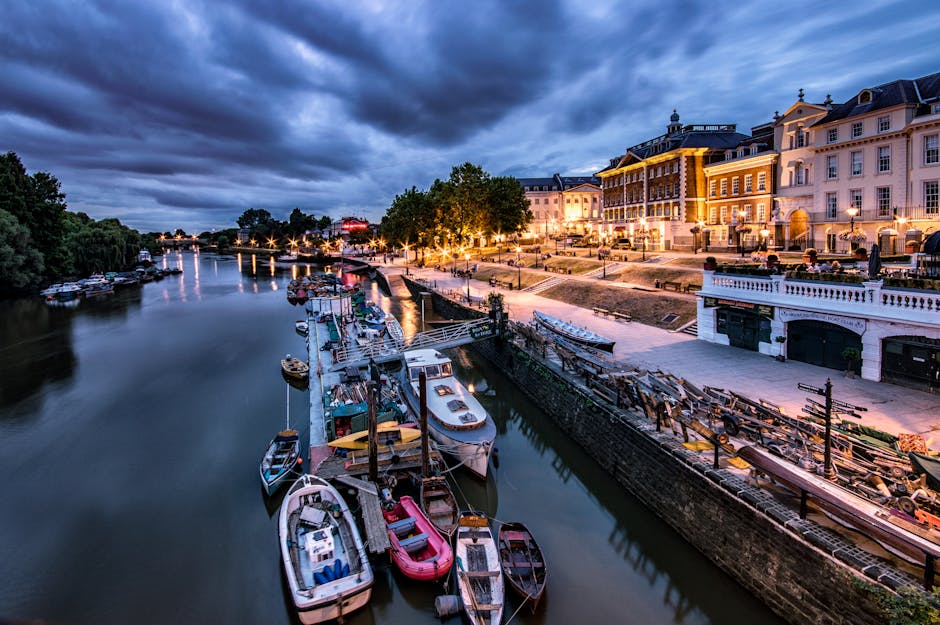 A tranquil riverside scene in the early evening featuring a collection of small boats docked along a narrow canal, with some boats covered by protective tarps and others exposed, showing a variety of colors including white, pink, black, and blue. The boats are moored to wooden piers and a stone canal wall, with some boats appearing to be fishing or leisure vessels. In the background, grand multi-storey buildings with classical architectural details, large windows, and decorative cornices are illuminated by warm exterior lighting, casting a gentle glow onto a paved promenade and nearby grassy areas with evenly spaced lampposts. The scene is set under a dramatic sky filled with dark, billowing clouds, reflecting a subtle hue from the evening light. To the left, lush green trees line the riverbank, contrasting with the historic buildings on the right. The overall atmosphere is calm, with the scene clearly connected to private or independent boat mooring and waterfront rubbish removal services often required for maintaining such areas in the context of waste management in urban waterways.