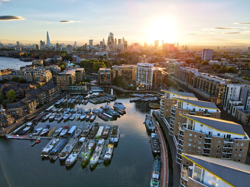 An aerial view of a cityscape during sunset, featuring a marina with numerous boats docked along the water's edge in the foreground. The marina is surrounded by modern residential buildings with balconies and flat rooftops, some with yellow accents along the edges. Beyond the marina, there are additional apartment blocks, trees, and streets leading towards a densely constructed urban skyline in the distance, which includes tall skyscrapers and iconic structures. The sky is illuminated with warm, golden hues, casting reflections on the water and creating a bright, atmospheric scene. The environment suggests a lively, waterfront residential area, with the scene captured in natural late-day light. This city scene exemplifies an urban environment where private landings and waterways are integrated with modern living spaces, supporting activities such as private boat mooring and on-site water-based leisure. The image aligns with the context of waste management services for dense city living areas, with visible structures indicating a well-developed, high-density environment typical for independent rubbish removal and waste disposal solutions.
