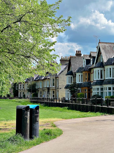 A row of Victorian-style terraced houses with bay windows, pitched roofs, and chimneys, situated along a residential street. In the foreground, there are two black waste bins placed on a grassy verge beside a paved pathway. A large tree with fresh green leaves extends its branches over the scene, partially shading the bins and sidewalk. Behind the houses, the sky is partly cloudy with patches of blue, suggesting a bright, partly sunny day. The image captures a typical suburban neighborhood scene, where private waste disposal solutions like the waste bins are positioned for curbside collection. The scene appears calm and well-maintained, with tidy front gardens and a traditional urban setting, fitting within the context of independent rubbish removal services typical for local homeowners.