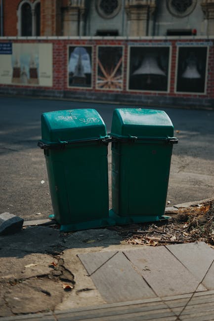 A row of Victorian-style terraced houses with bay windows, pitched roofs, and chimneys, situated along a residential street. In the foreground, there are two black waste bins placed on a grassy verge beside a paved pathway. A large tree with fresh green leaves extends its branches over the scene, partially shading the bins and sidewalk. Behind the houses, the sky is partly cloudy with patches of blue, suggesting a bright, partly sunny day. The image captures a typical suburban neighborhood scene, where private waste disposal solutions like the waste bins are positioned for curbside collection. The scene appears calm and well-maintained, with tidy front gardens and a traditional urban setting, fitting within the context of independent rubbish removal services typical for local homeowners.