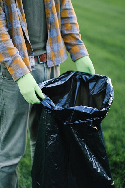 A person standing outdoors on a grassy area, wearing a plaid shirt with shades of yellow, gray, and blue, along with green rubber gloves, is holding open a large black rubbish collection bag made of glossy plastic material. The bag is partially filled, with visible creases and an empty interior, ready for waste disposal. The person's lower half includes light-colored trousers and a brown belt. The background features a blurred view of well-maintained green grass and natural daylight, emphasizing an environment suitable for waste collection or garden clearance activities often associated with private rubbish removal services. The scene highlights a methodical approach to waste management involving independent collection, aligning with practices for seasonal garden waste removal or off-site rubbish clearance.