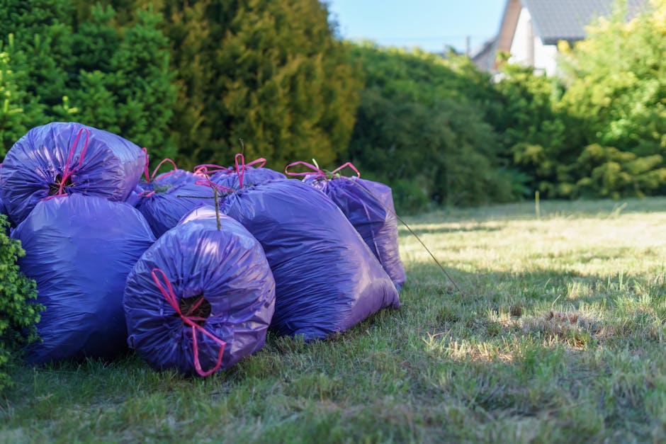 A group of seven large purple rubbish bags made of crinkled plastic material, each secured with pink drawstrings, are stacked and arranged along the edge of a grassy area. The bags appear full, with some slightly bulging, and are placed on a patch of natural green grass with a few uneven patches. In the background, dense, leafy green trees and shrubs form a natural boundary, with some taller trees standing behind the bushes. Partially visible at the top right are residential buildings with grey roofs, indicating a suburban environment. The scene is illuminated by natural daylight, casting soft shadows on the grass, and the overall setting suggests an outdoor clearance operation, possibly as part of a private rubbish removal or garden waste collection service, such as those offered by companies like rubbishremovalkingstonuponthames.co.uk, supporting a seasonal garden waste clearance context.