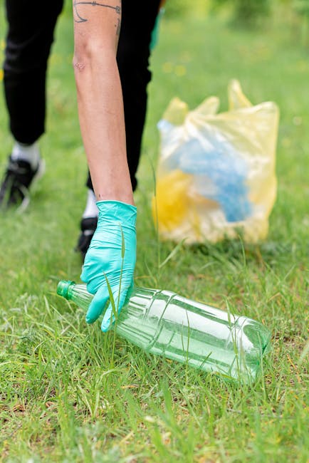 A person standing outdoors on a grassy area, wearing a plaid shirt with shades of yellow, gray, and blue, along with green rubber gloves, is holding open a large black rubbish collection bag made of glossy plastic material. The bag is partially filled, with visible creases and an empty interior, ready for waste disposal. The person's lower half includes light-colored trousers and a brown belt. The background features a blurred view of well-maintained green grass and natural daylight, emphasizing an environment suitable for waste collection or garden clearance activities often associated with private rubbish removal services. The scene highlights a methodical approach to waste management involving independent collection, aligning with practices for seasonal garden waste removal or off-site rubbish clearance.
