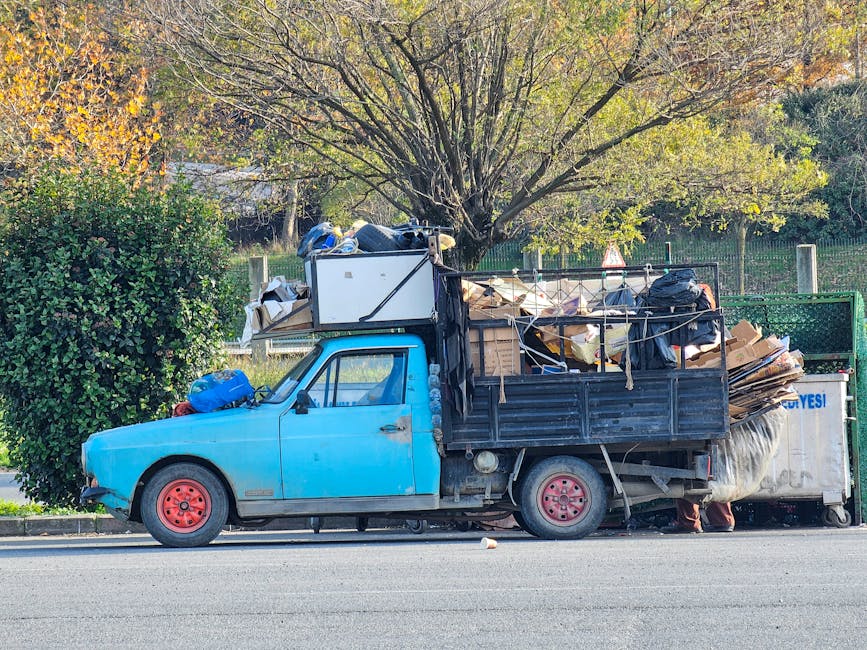 A small blue utility truck parked on the side of a street, heavily loaded with miscellaneous rubbish and debris in its open-top cargo area. The vehicle's body shows signs of age with visible rust spots and uneven paint, particularly around the front wheel arch and door. The cargo area is filled with various materials, including cardboard boxes, bags, and wooden planks, which are piled high and secured with ropes. A white refrigerator is positioned on top of the rubbish, extending beyond the truck's height, with its doors taped shut. The tires are pinkish-red, and the rear wheel on the driver's side appears slightly deflated. Behind the truck, there is a lush green hedge and a large tree with a full canopy of green leaves turning yellow and orange, indicating autumn. The background includes a green metal fence and a glimpse of a park or garden area, suggesting the vehicle is parked near a residential or suburban setting. The overall scene reflects private waste handling, typical of independent rubbish collection or on-site clearance services, with natural lighting indicating a calm daytime environment.