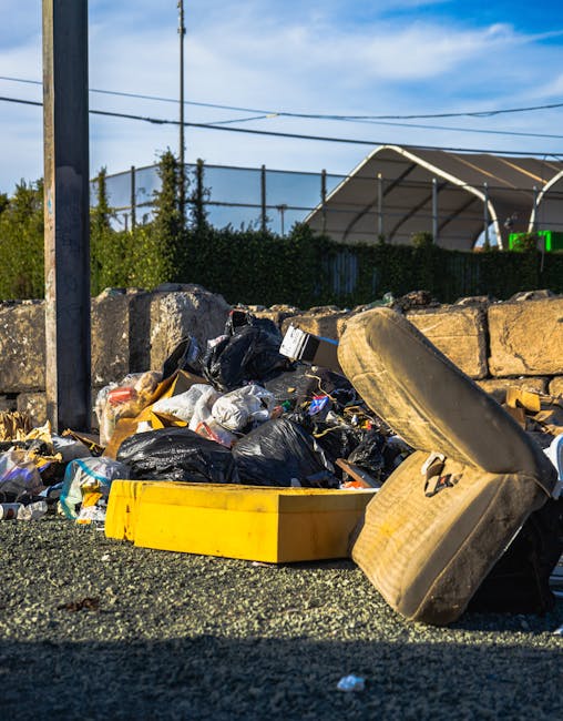 A collection of dumped waste on the side of a paved area shows various materials including black garbage bags, a yellow plastic container, a worn car tire, and scattered debris. The waste is positioned against a low stone wall composed of large, uneven beige stones. In the background, there is a lattice fence with dense green foliage and trees beyond, along with an outdoor sports court enclosed by a high metal fence, partially visible under a blue sky with some wispy clouds. To the left, a metal pole is visible, possibly supporting electrical wires which span across the scene. The environment appears to be an area designated for rubbish accumulation, possibly reflecting improper disposal or an onsite collection point. The scene is lit by natural daylight, emphasizing the different textures of the plastic, rubber, stone, and asphalt, fitting within a context of private waste handling and waste removal services offered by companies like rubbishremovalkingstonuponthames.co.uk, which may be involved in managing such waste pickups.